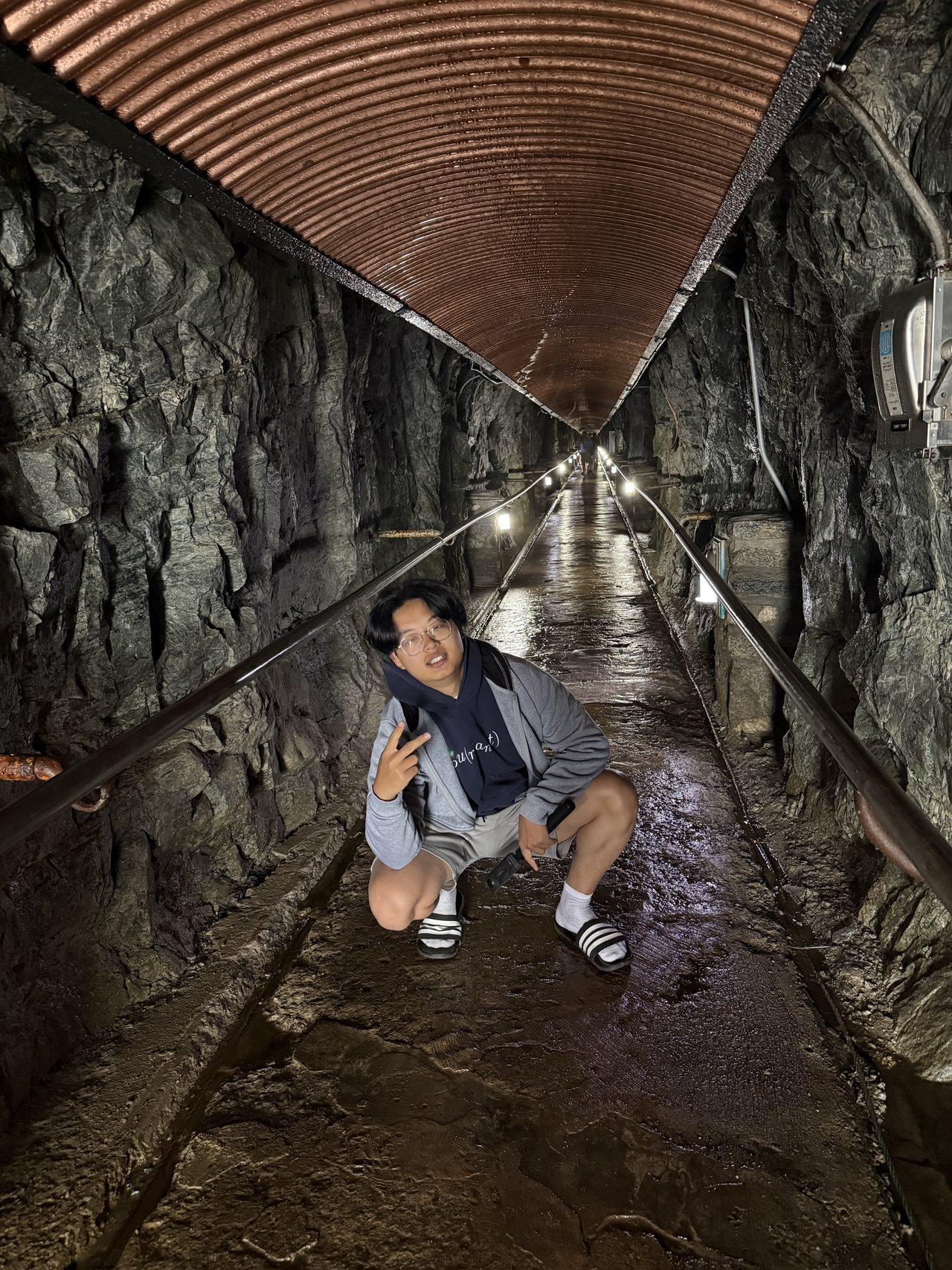 A long rock tunnel with a copper corrugated ceiling and strung lights.