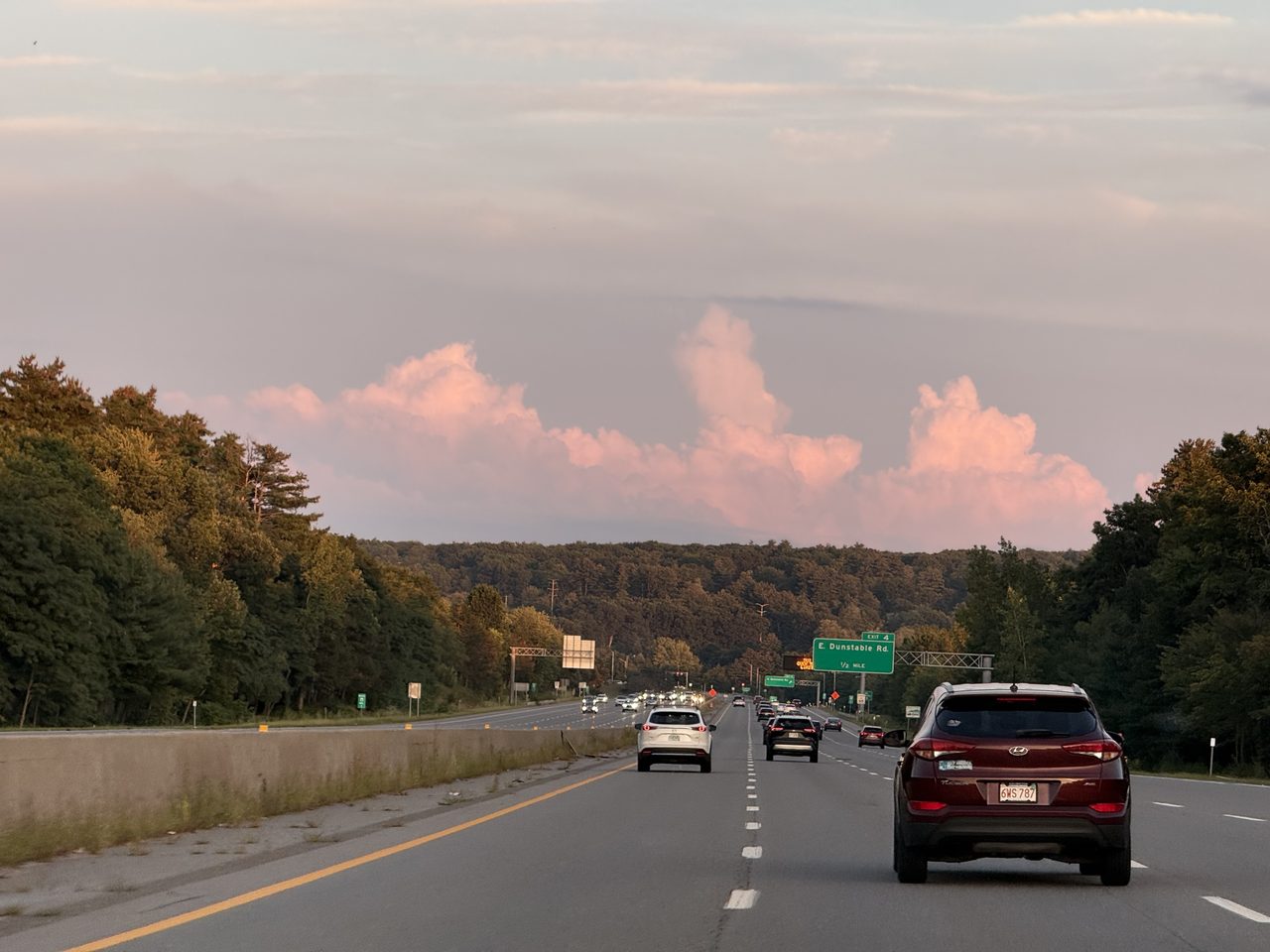 Pink-lit cumulus clouds over an evening highway with an exit sign for E. Dunstable Road.