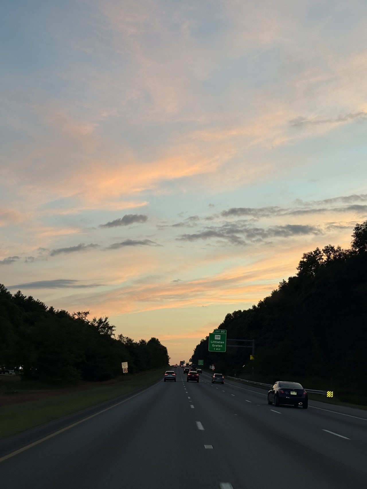 An evening highway under an orange and pink sunset sky near an exit sign for Littleton and Groton.