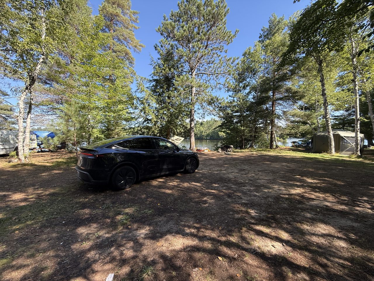 A black Tesla parked under pines beside a lake with a tent nearby.