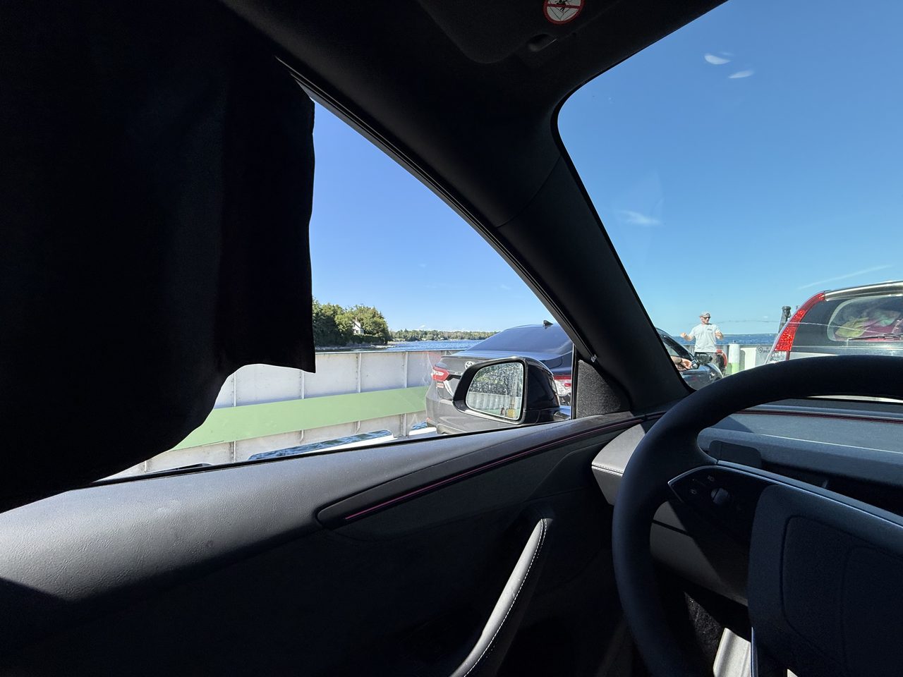 View from inside a car boarding a small vehicle ferry, water and shoreline ahead.