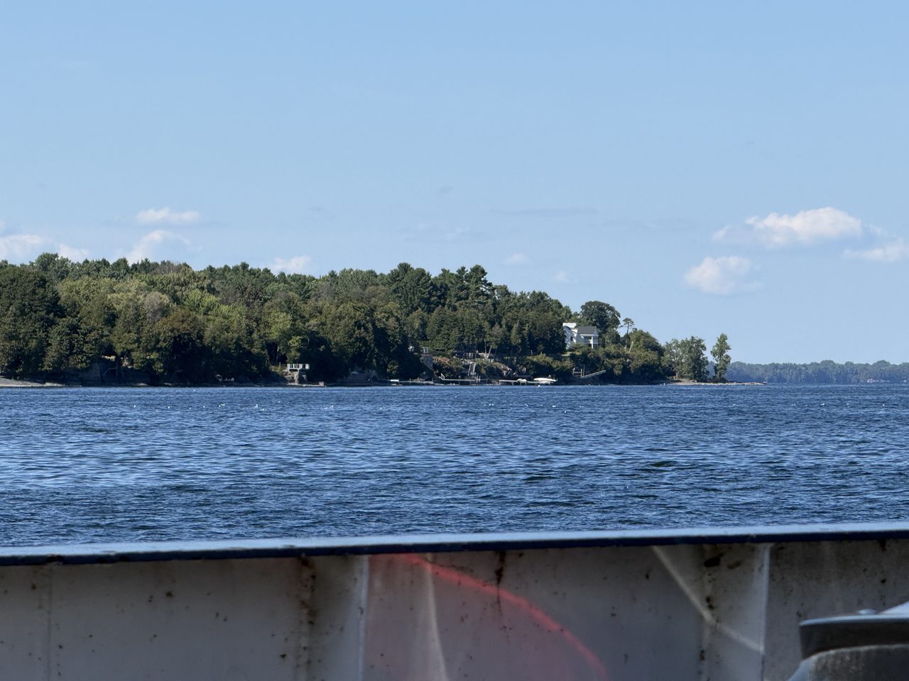 Lake Champlain and a wooded shoreline seen over a ferry's white railing.