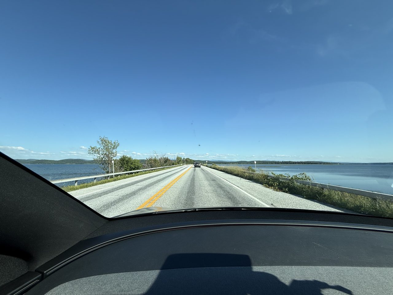A roadside guardrail with lake water and distant hills beyond trees.