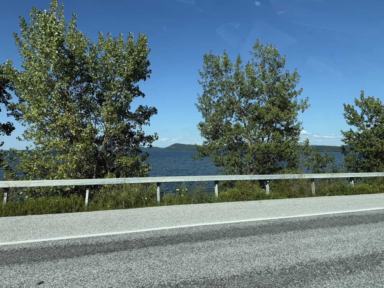 Lake water seen past a highway guardrail and roadside trees.