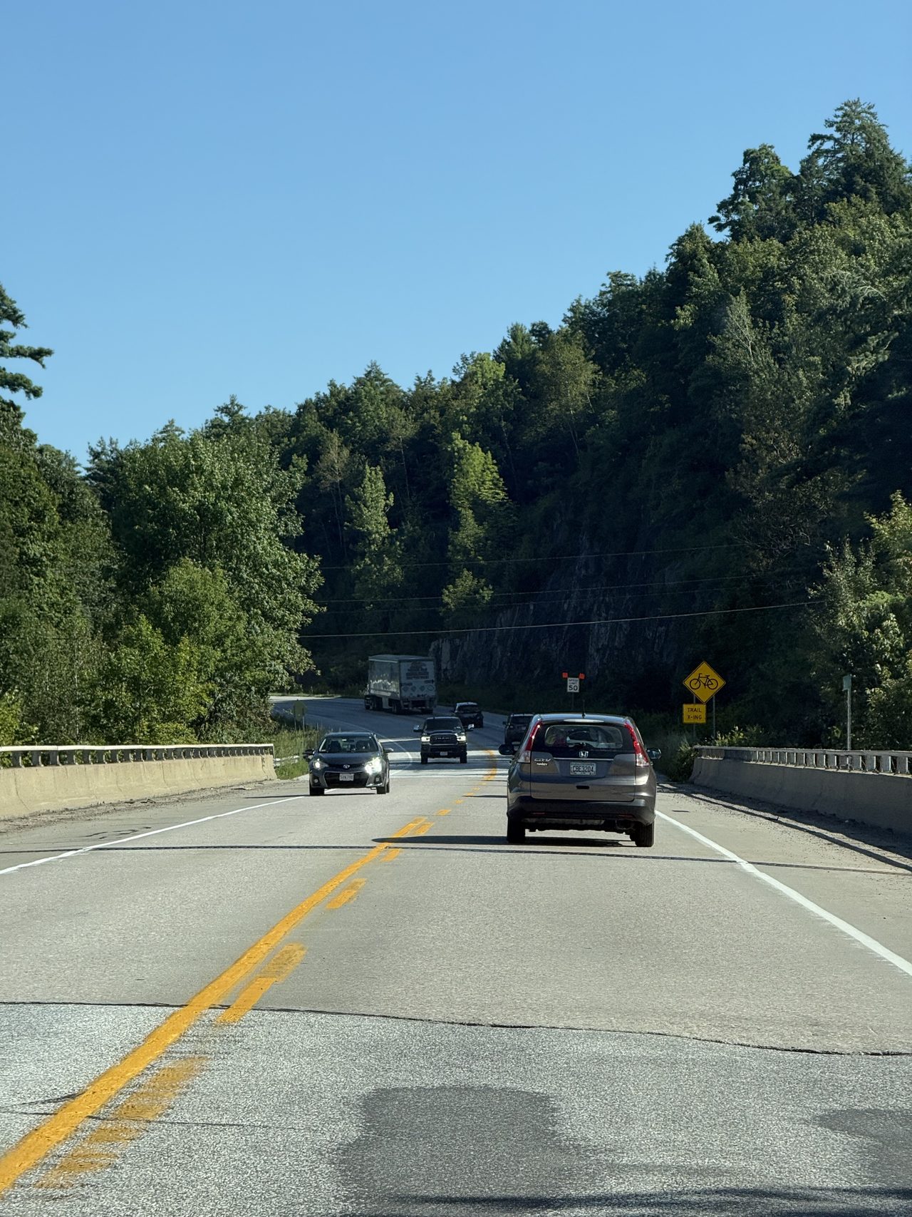 A highway between forested hills with a rock cut ahead.