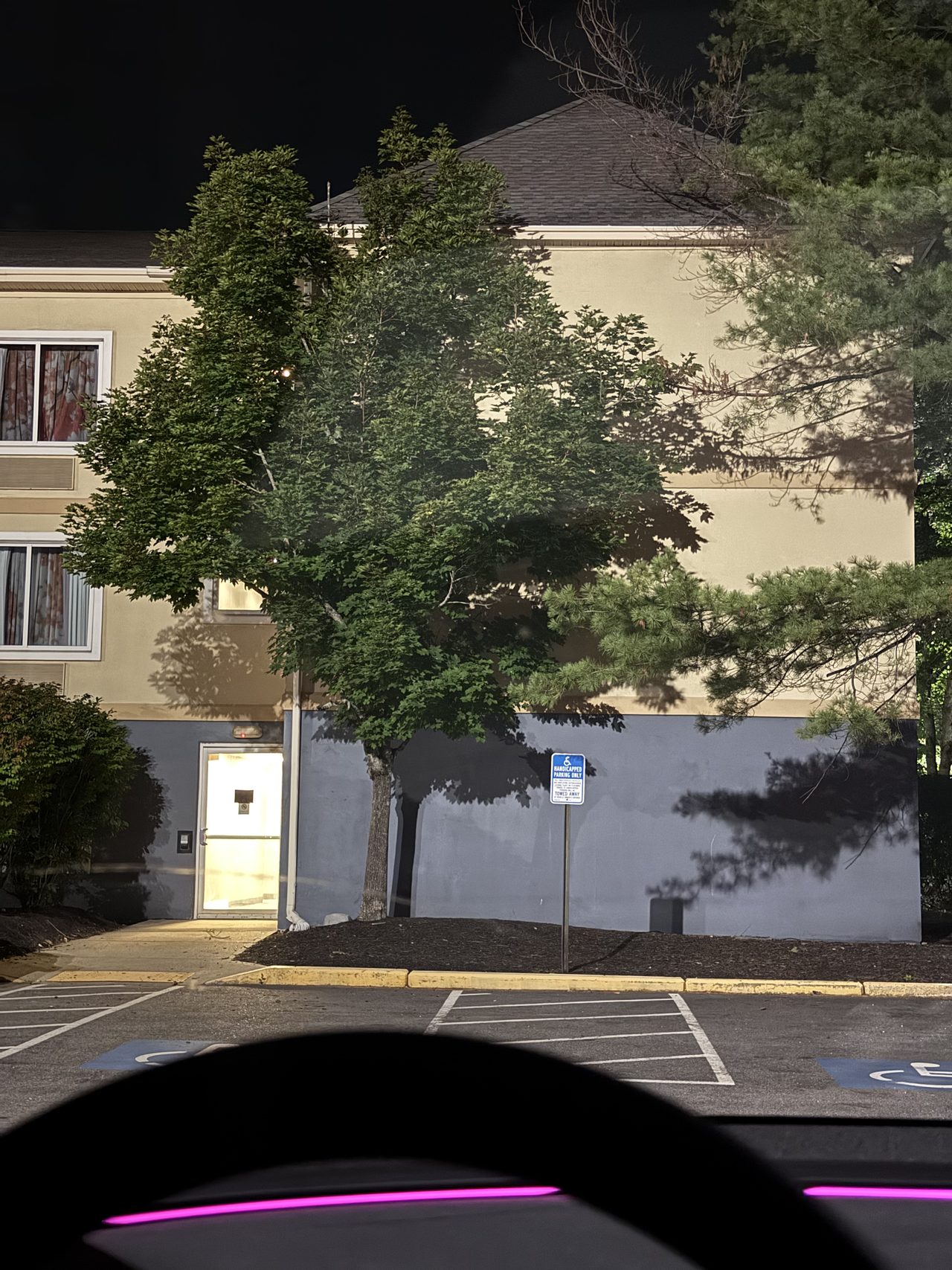 A motel building at night with trees and a lit doorway seen from a parking lot.