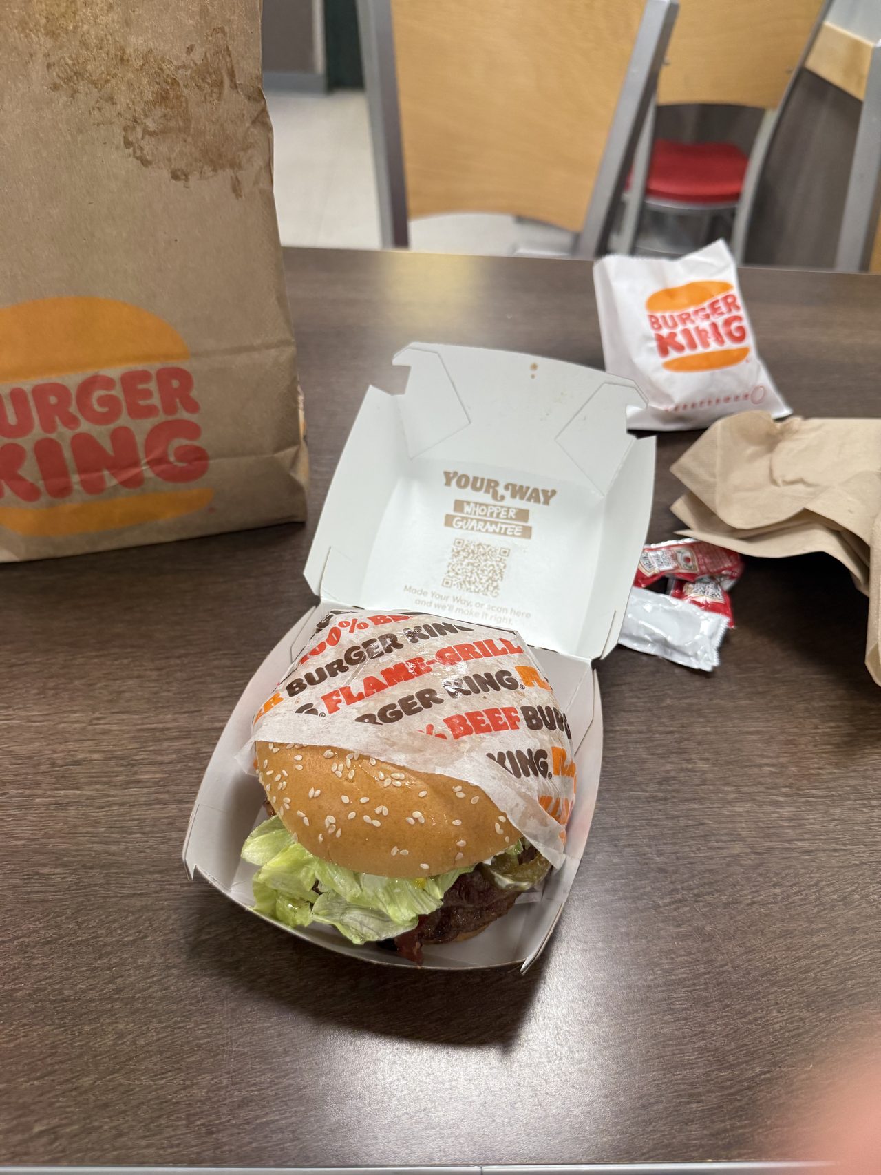 A Burger King meal on a table with bag, napkins and ketchup packets in an empty restaurant.