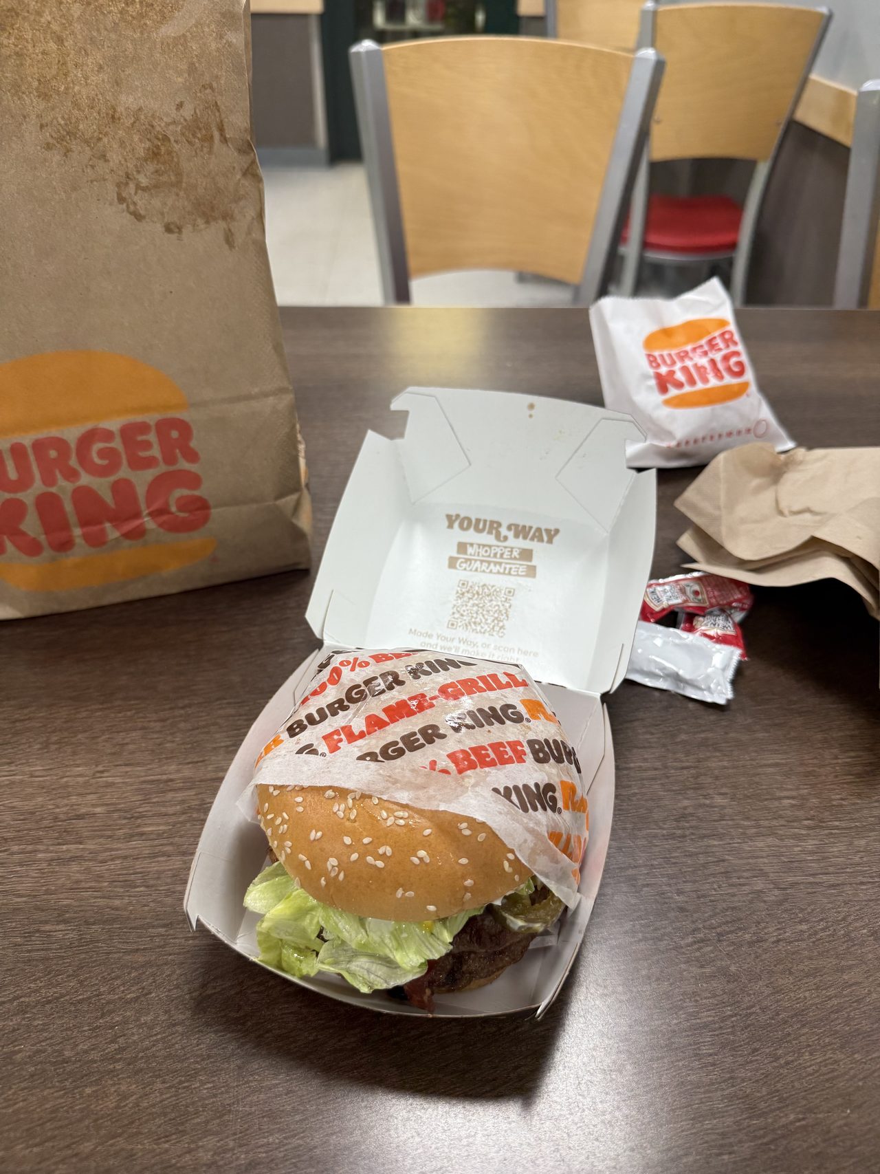 A Whopper in an open Burger King box on a table with a paper bag and ketchup packets.