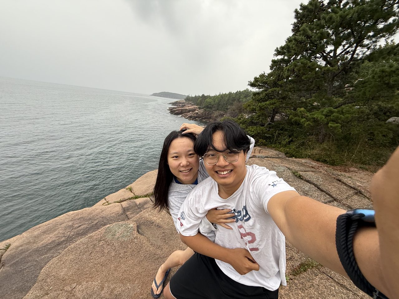A couple's wide selfie on granite shore with a misty cove and evergreens behind.