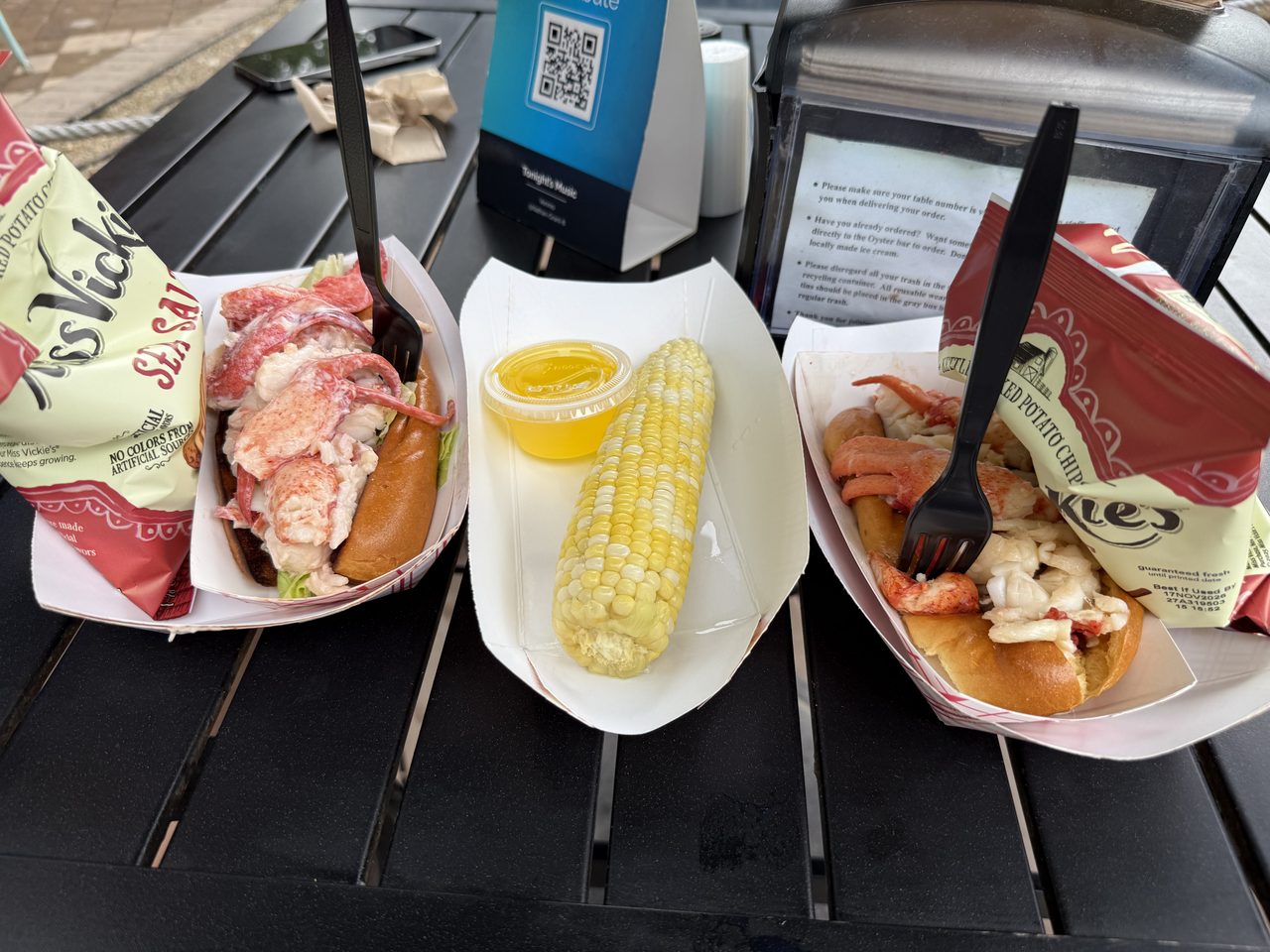 Two lobster rolls in paper trays with corn on the cob and a cup of butter.