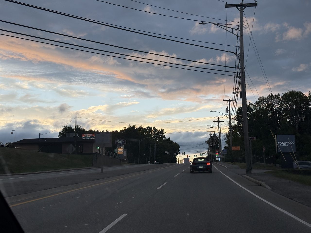 A car following a Mustang on a town road at dusk with orange-lit clouds.