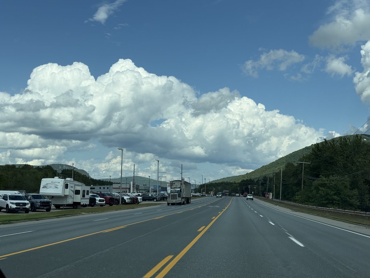 A four-lane road with towering white cumulus clouds over green hills.