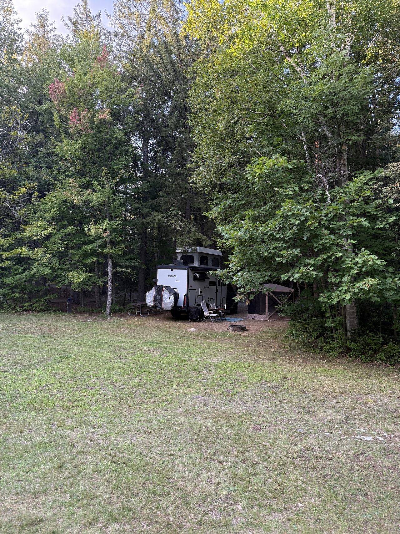 A camper and screen tent at the edge of a forest clearing with mown grass in front.