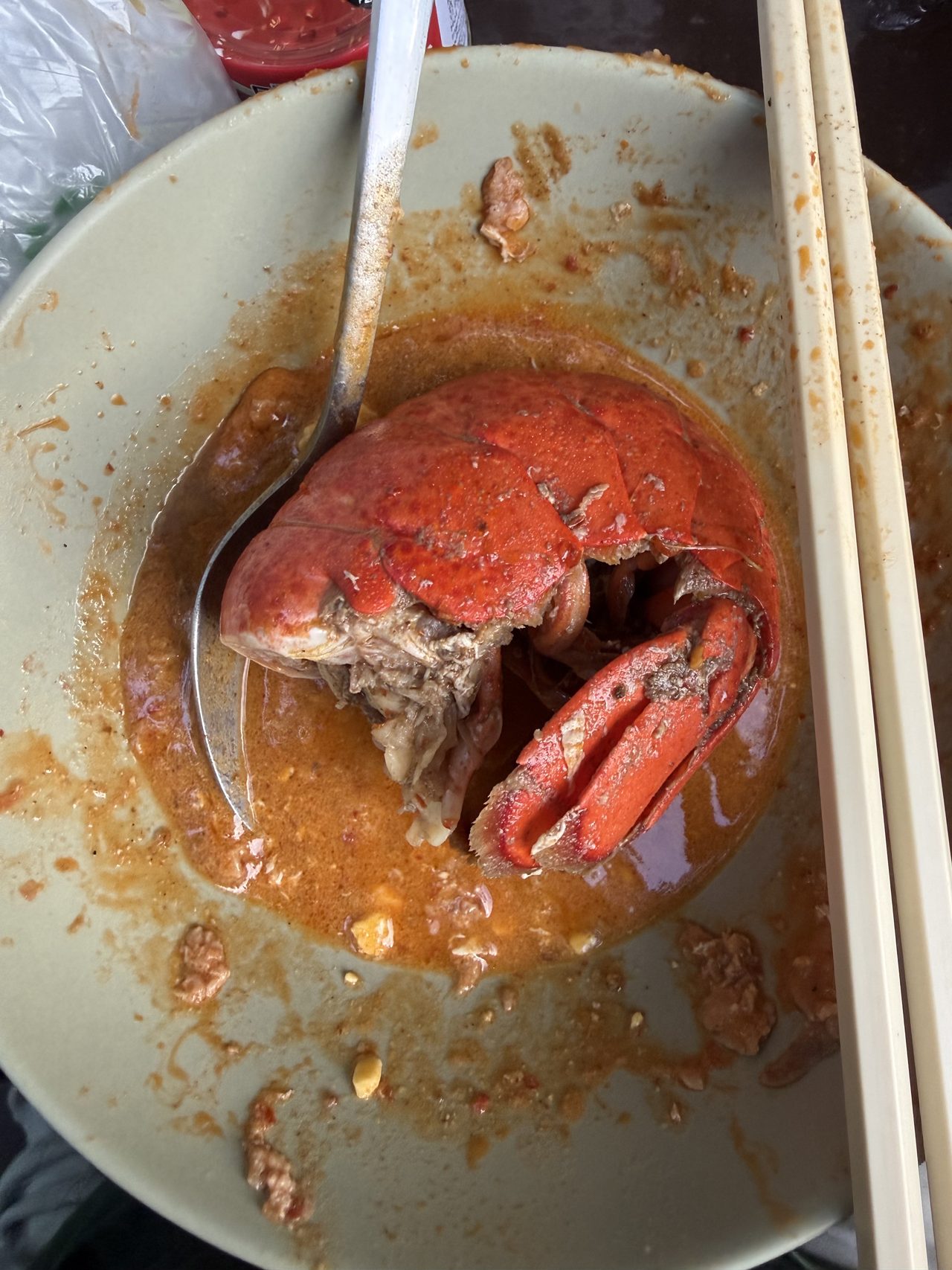A cooked lobster head and claw sitting in a bowl of red hotpot broth with chopsticks.