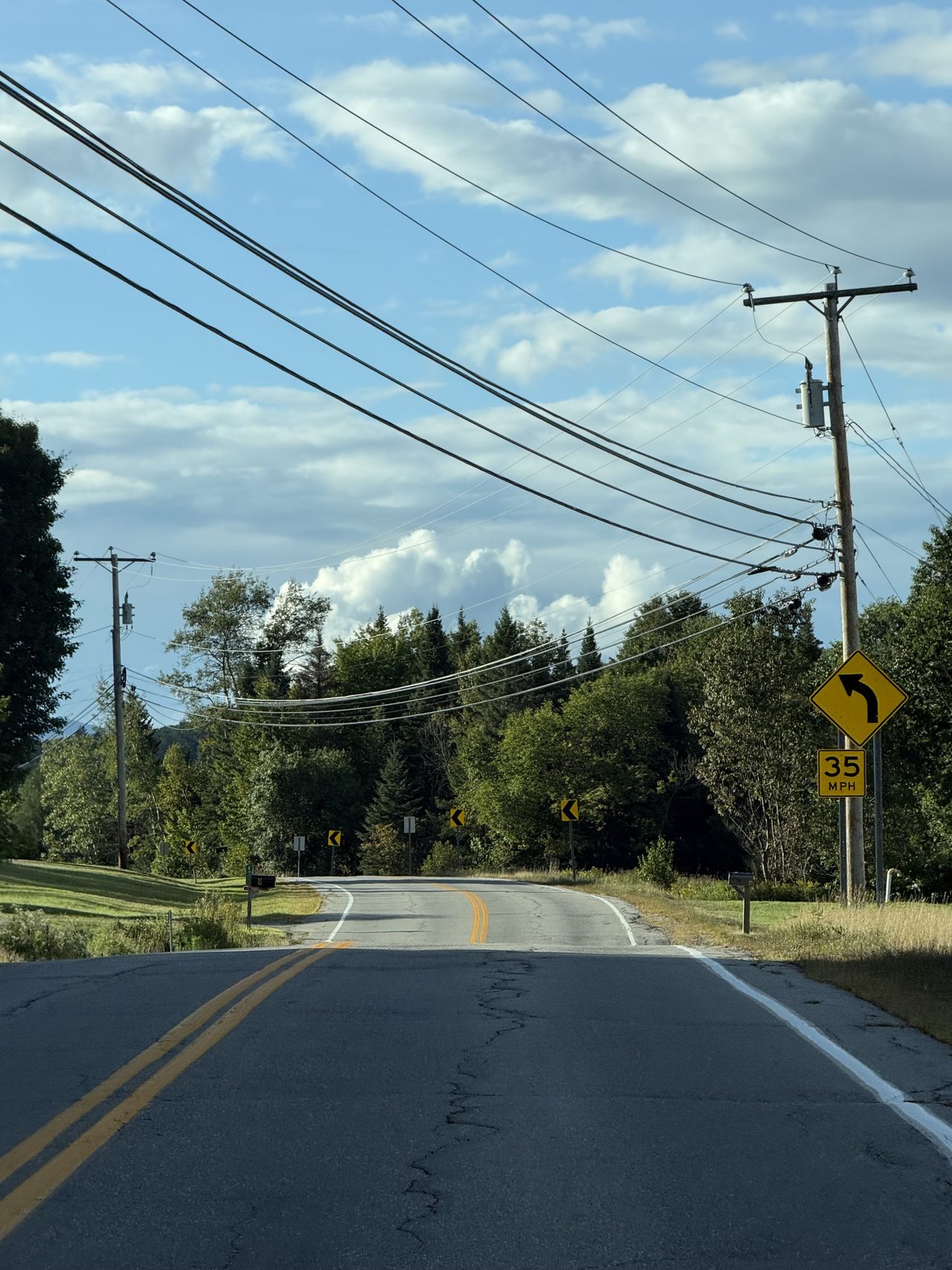 An empty two-lane road curving through trees under a blue sky with power lines.