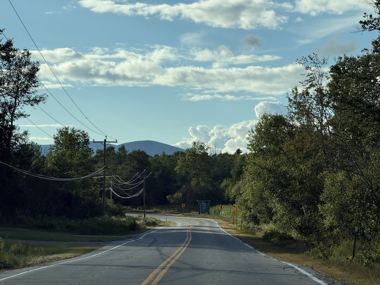 A curving country road with a blue mountain in the distance under scattered clouds.