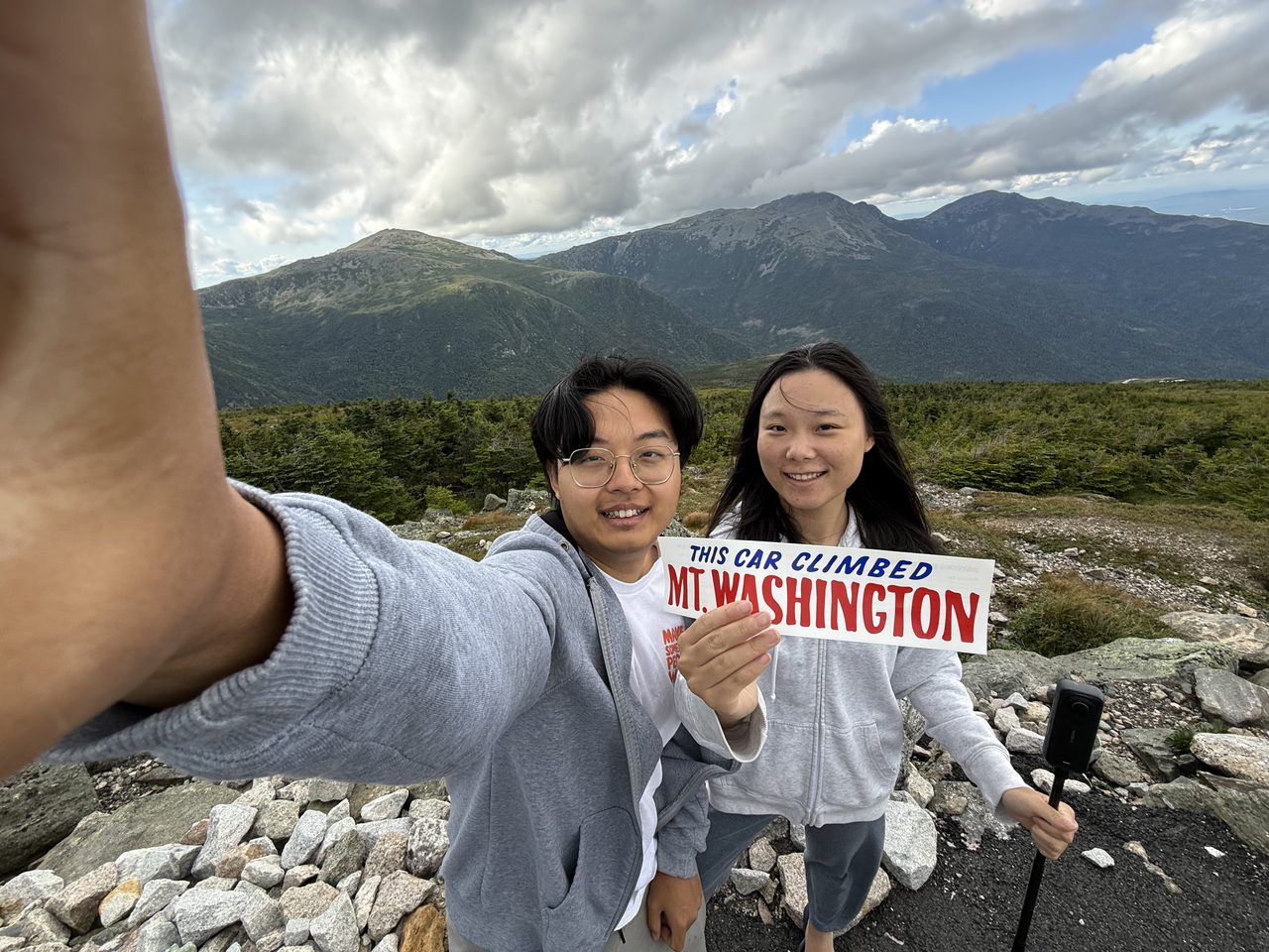 Two people taking a selfie holding a bumper sticker reading This Car Climbed Mt. Washington, mountains behind.