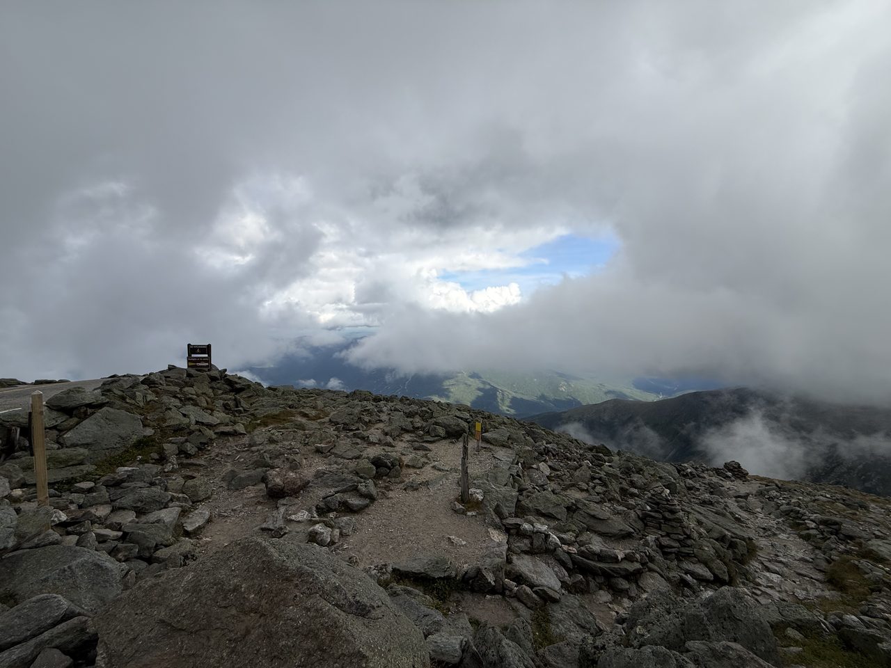 A rocky summit area with a trail sign, mostly enveloped in low cloud with one patch of blue.