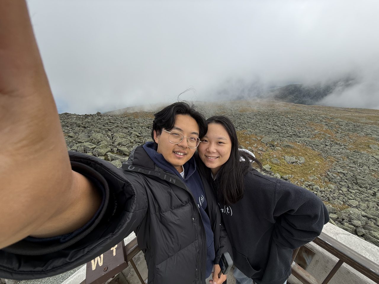 A couple in winter jackets taking a selfie on a boulder-covered summit in cloud.