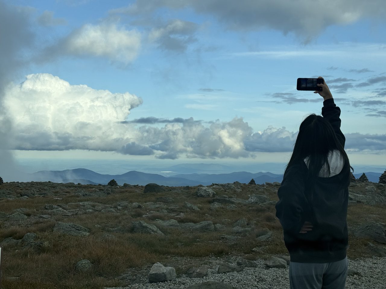 A person photographing a vast view of ridges and distant lakes from an alpine slope.