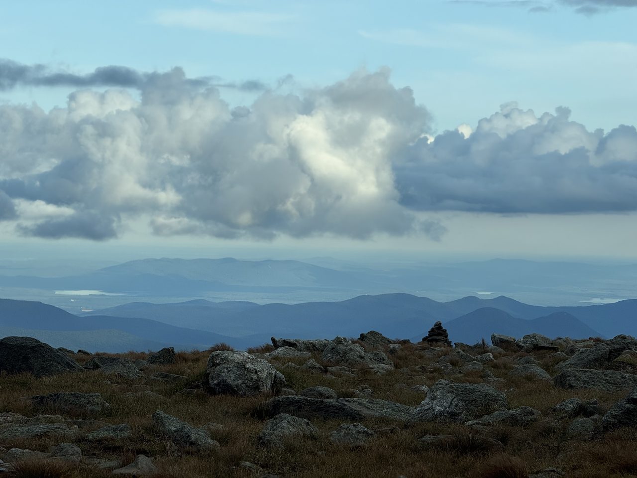 A stone cairn on alpine tundra with layered blue ridges and distant lakes beyond.