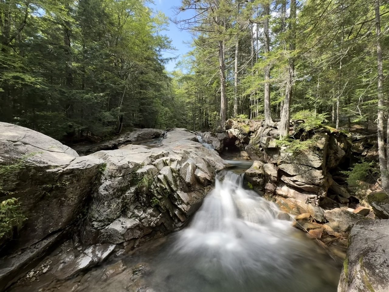 A stream cascading over granite ledges in bright sunlight with blue sky above.