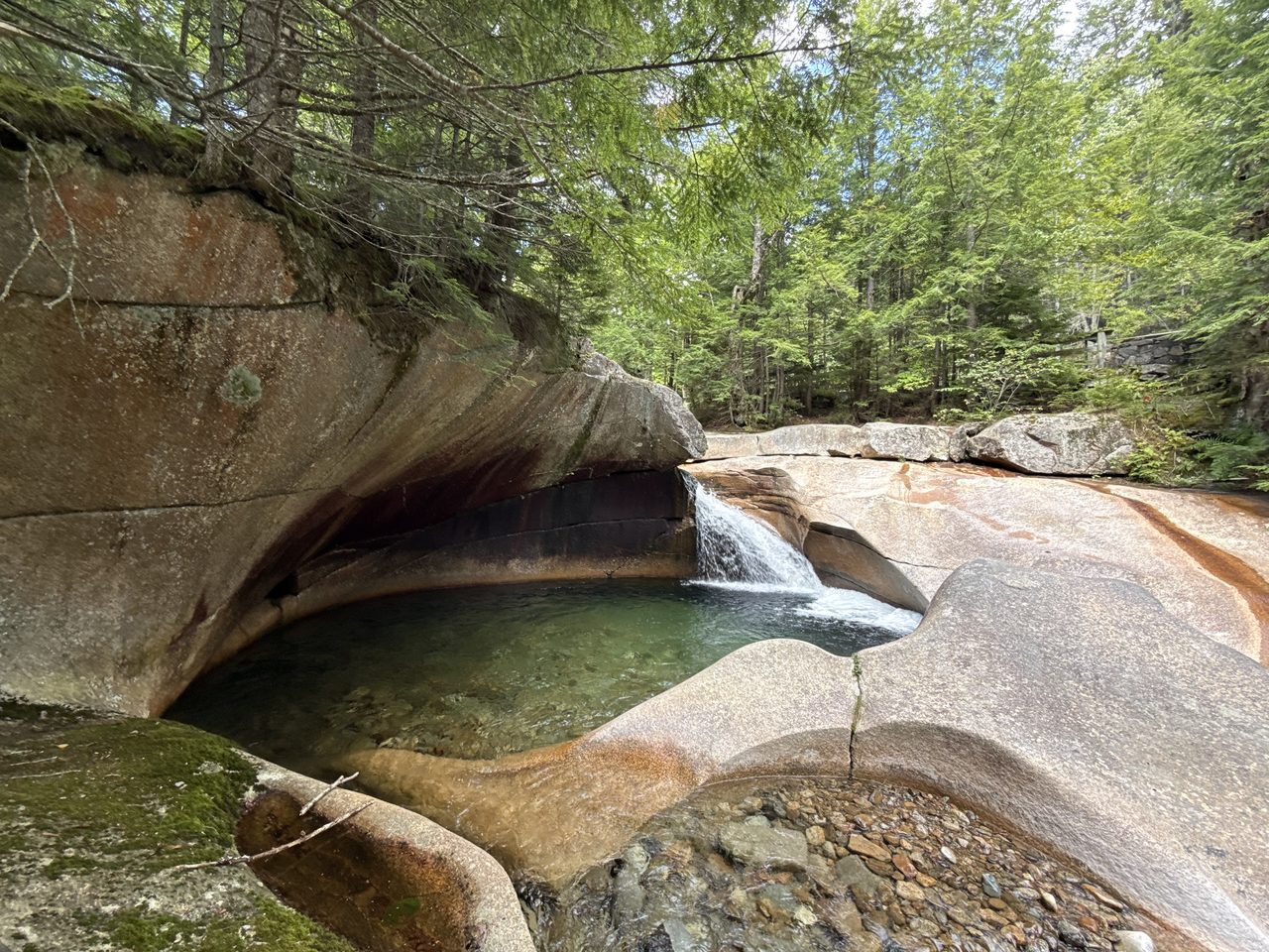 A large circular granite pothole filled with clear green water and a small waterfall.